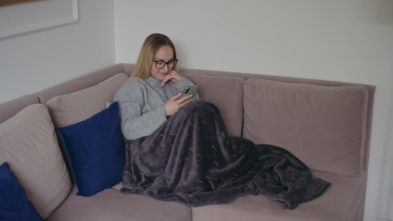 Una giovane donna sorridente con gli occhiali è seduta comodamente su un divano, avvolta in una morbida coperta, mentre usa il suo smartphone. Appare rilassata e felice in un accogliente e moderno ambiente di soggiorno.