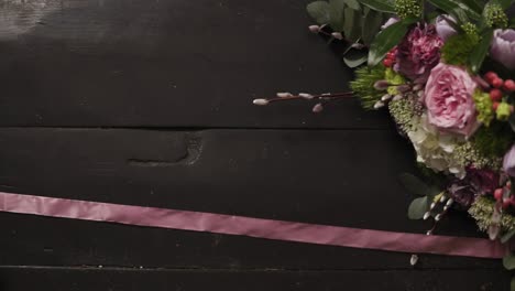 Two-silk-pink-ribbons-sprawled-on-the-dark-wooden-surface,-a-beautiful-bouquet-of-flowers-in-a-frame.-Slow-motion