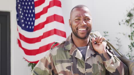 portrait of happy african american male soldier holding bag with flag of usa in the background