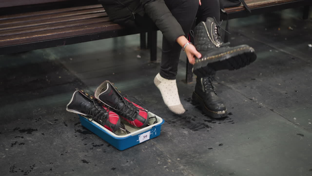 Female sitting on bench offing black leather boots with zippers while red and black ice skates rest inside blue plastic tray on wet scratched floor during preparation for winter skating activity