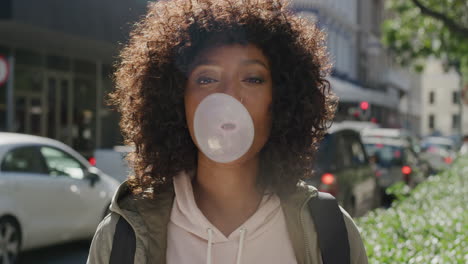 retrato de una joven mujer afroamericana soplando chicle sonriendo feliz disfrutando de una diversión juguetona en la vibrante calle de la ciudad por la mañana estudiante con peinado afro