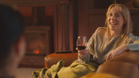 two female friends relaxing and laughing on sofa in lounge with cosy fire holding glass of wine