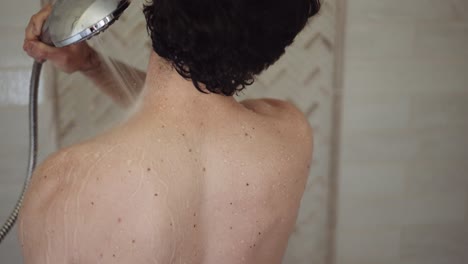 white man with dark hair washes his back and neck with the shower head in his hand