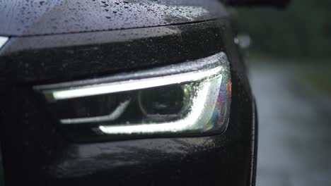 black high end car led headlight turning on in the rain as dew drops form on the hood, extreme close up