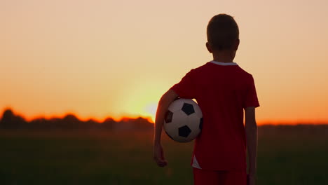 un niño de pie en el campo al atardecer con una pelota de fútbol mira en la distancia y sueña con convertirse en un exitoso jugador de fútbol. pararse y ver el sol ponerse en el campo