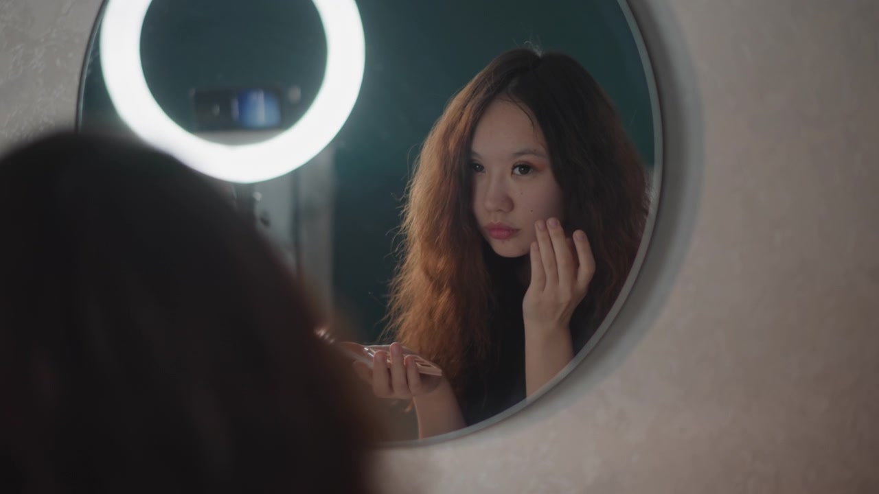 Una giovane donna con capelli rossi ricci si trucca davanti a uno specchio rotondo illuminato da una ring light, tenendo il rossetto vicino alle labbra mentre controlla il suo aspetto in una tranquilla routine di bellezza al chiuso e con luce soffusa.
