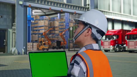 close up back view of asian male engineer with safety helmet working on a green screen laptop and looking around while standing , outside of logistics distributions warehouse