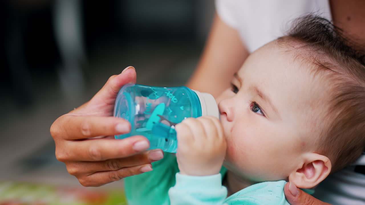 Lovely toddler with stylish hair drinking water from a bottle. Mom's hand is holding a bottle for her kid. Close up.