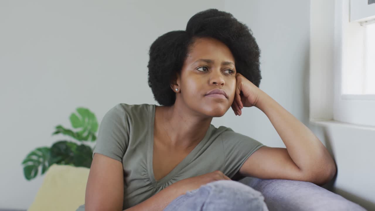 Thoughtful african american woman sitting on sofa, looking through window