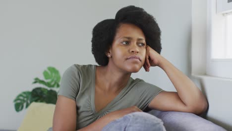 Thoughtful-african-american-woman-sitting-on-sofa,-looking-through-window