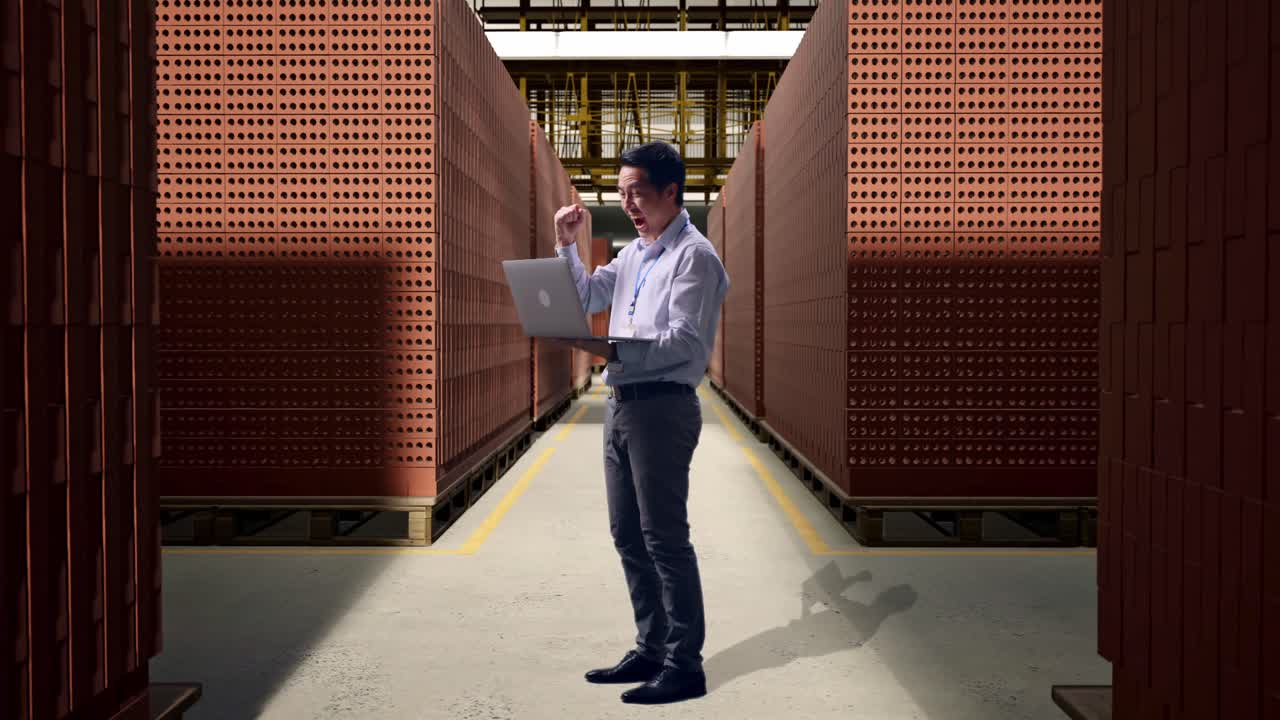 Full Body Side View Of An Asian Male Professional Worker Standing With His Laptop With Red Brick Packed in Stacks Are Stored, He Raises His Fist Up With Screaming Goal After Typed On The Laptop