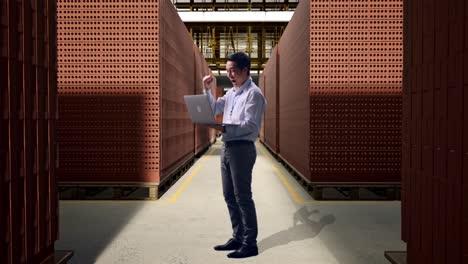 full body side view of an asian male professional worker standing with his laptop with red brick packed in stacks are stored, he raises his fist up with screaming goal after typed on the laptop