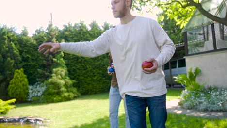 zijaanzicht van een blanke jongeman die op een zonnige dag een rode petanquebal in het park gooit terwijl zijn vrienden op hun beurt wachten