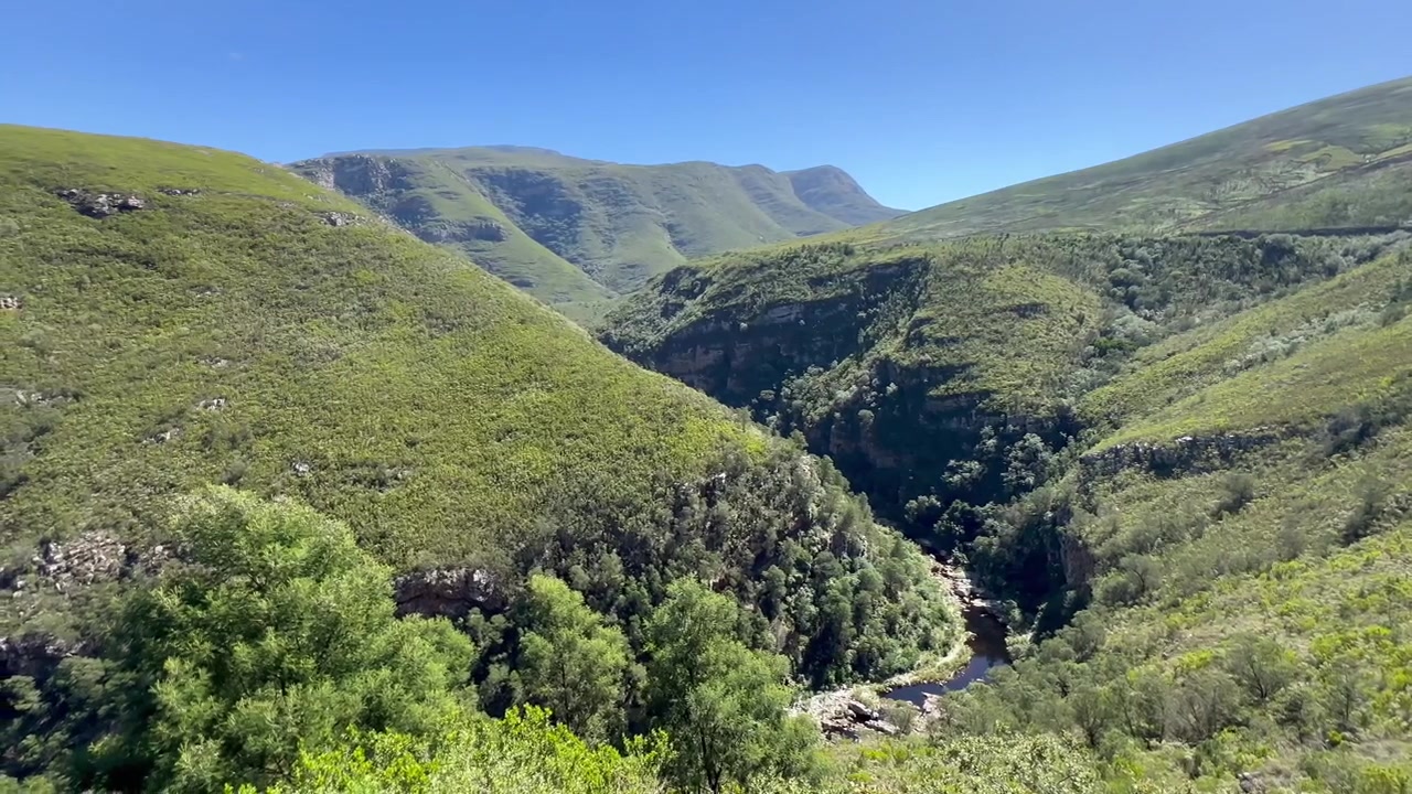 Valle di un verde lussureggiante con un fiume che scorre attraverso un terreno montuoso sotto un cielo azzurro limpido a Riversdale, Sudafrica.