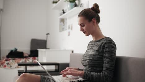 Side-view-of-young-woman-at-home-sitting-on-the-sofa,-working-with-a-laptop-and-typing-text-fast-looking-at-the-screen