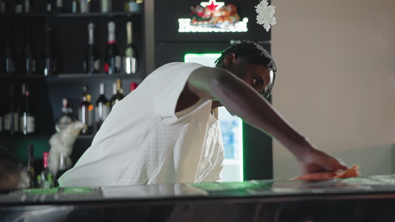 Bartender focused on cleaning and setting up bar counter. Shelves stocked with bottles and festive Heineken display. A rabbit sits on the counter, adding an unexpected touch to the scene