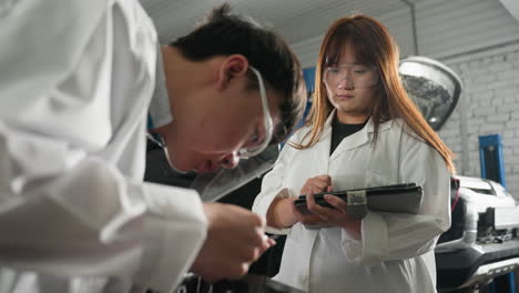 student in mechanical workshop wearing protective goggles, measuring engine dimensions with vernier caliper, female colleague nearby operates tablet, recording data. industrial setting with tools