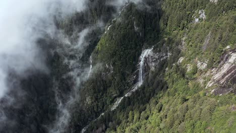 drone aerial view of a waterfall and a torrent on the side of a mountain, big cloud hiding the forest underneath in the shadow in northern italy