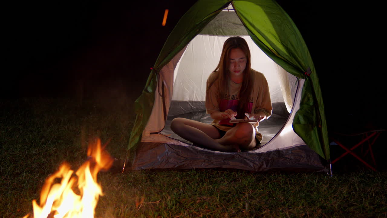 아시아 여성이 야외 캠핑을 하고 있으며, 텐트 안에서 5g로 연결된 현대 스마트폰으로 불 앞에서 채팅을 하고 있다.