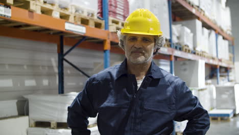 trabajador senior feliz en casco de pie en el almacén y mirando a la cámara con los brazos cruzados