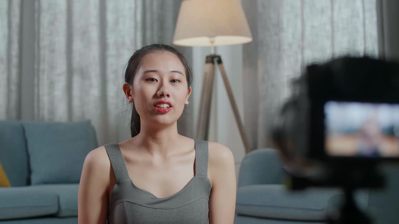 joven entrenadora asiática mujer en ropa deportiva hablando a la cámara y haciendo hombro-brazo cruzado mientras graba ejercicio de enseñanza en casa