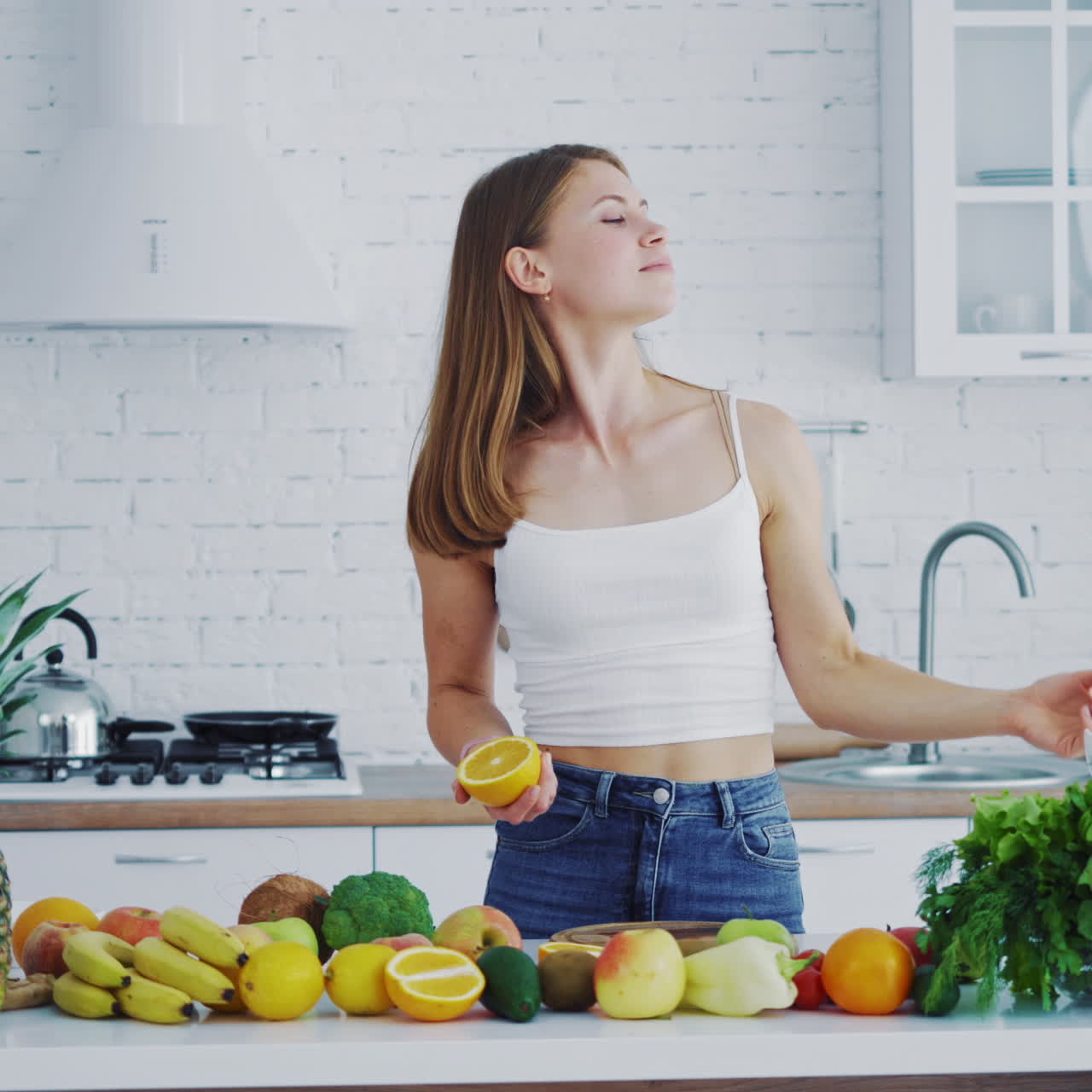Beautiful female in the light kitchen. Smiling young woman proposing fresh orange. Healthy organic fruit and vegetables on the table.