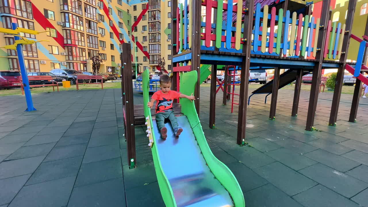 Caucasian toddler in bright t-shirt and pants playing on the playground. Kid slides to the ground and runs away.