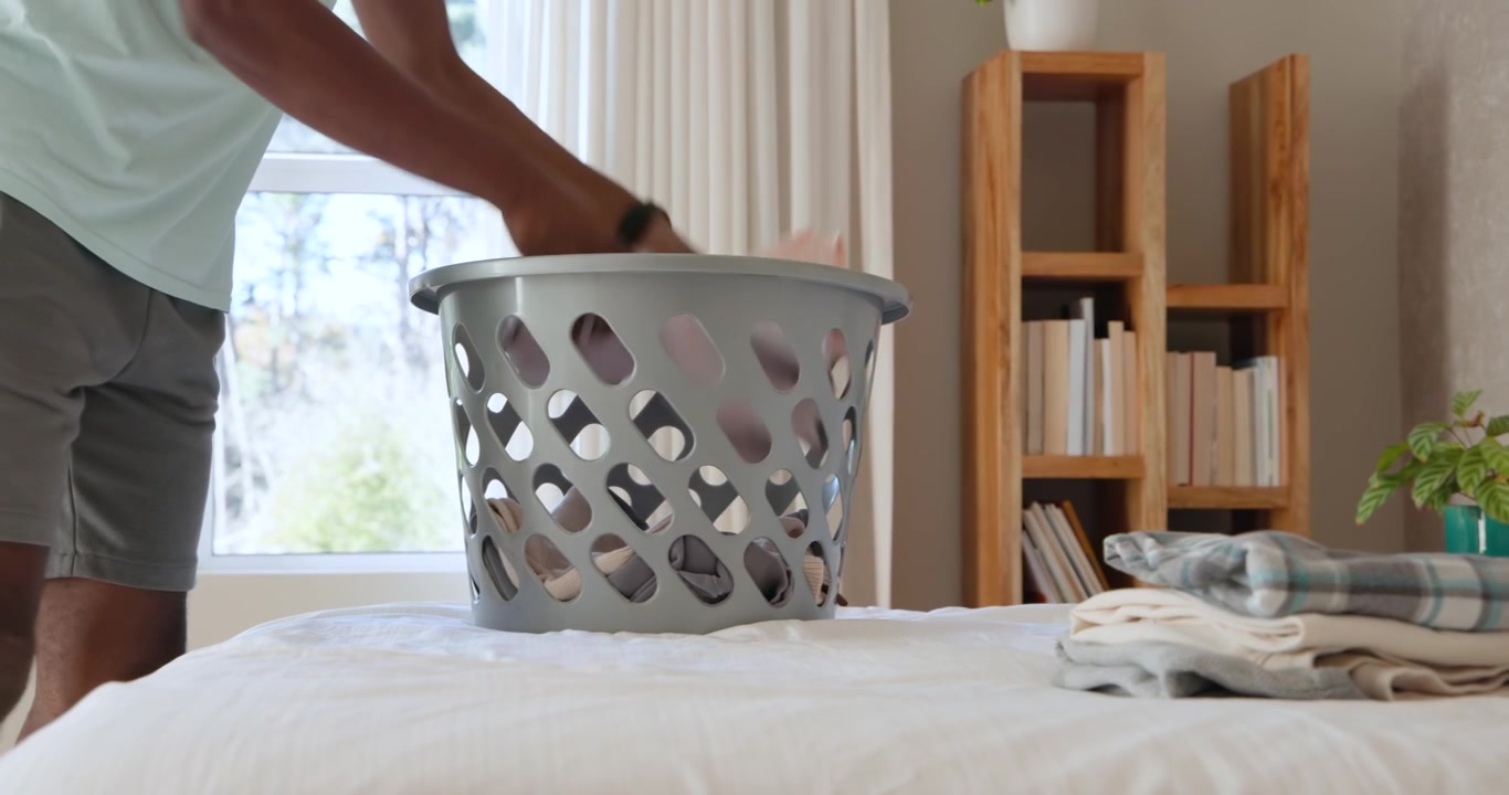 Person entering from left is picking folded garments from bed and loading gray basket for tidying