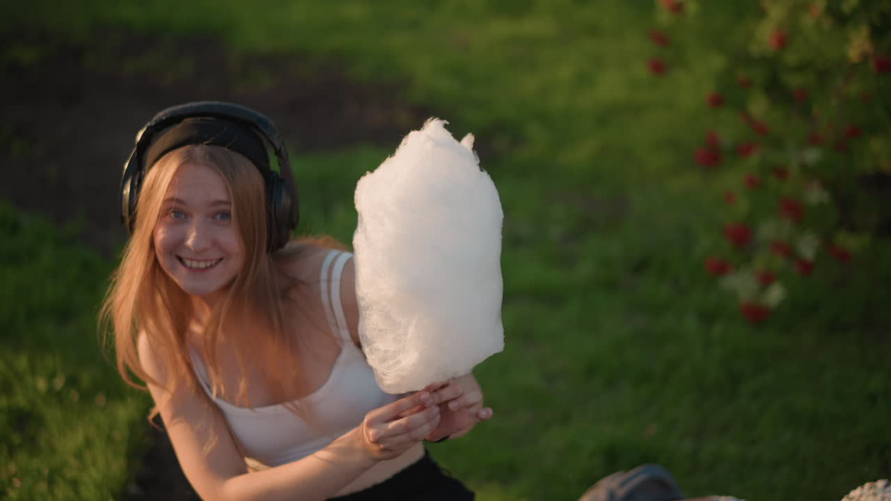 Giovane donna felice che si nasconde dietro caramelle, ragazza bionda allegra che ridacchia con le cuffie tra le dolcezze, ambiente luminoso del parco che cattura una ragazza innocente e giocosa con delizie zuccherine e musica
