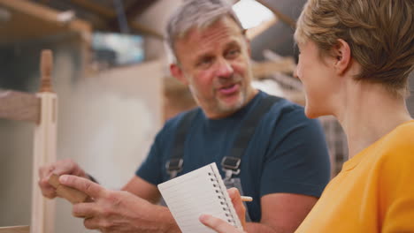 female apprentice making notes learning from mature male carpenter in furniture workshop