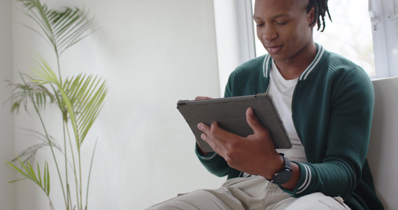 Homme afro-américain d'une vingtaine d'années en cardigan vert, tenant une tablette, tapant sur l'écran en lisant près d'une fenêtre