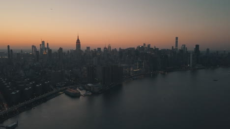 AERIAL:-Over-East-River-showing-Manhattan-New-York-City-Skyline-in-Beautiful-Dawn-Sunset-Orange-Light-right-before-Dark
