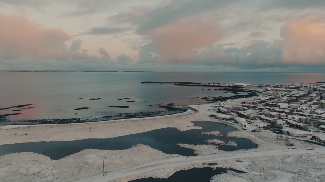 Letecký pohled z dronu na malé pobřežní městečko na Islandu pokryté sněhem v zimě. Odlehlá severská osada obklopená zamrzlým terénem, oceánem a dramatickou oblohou při západu slunce.