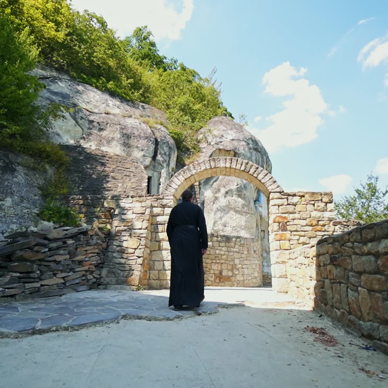 Monk wearing long black robe goes by the territory of rock monastery. Following the priest of orthodox church. Low angle view