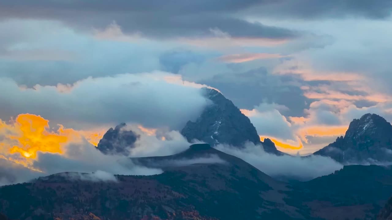 Moody sunrise timelapse over Grand Teton and Table Mountain as low clouds drift across rugged peaks, revealing warm alpenglow and layered mountain scenery
