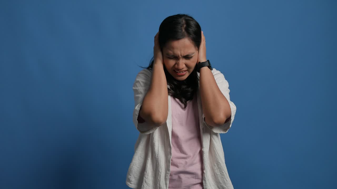 Unhappy sad angry young asian woman close eyes and cover ears by hands being annoyed with loud noise, isolated blue studio background, emotions, dislike loud sounds