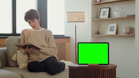 woman reading a book on a sofa with a tablet