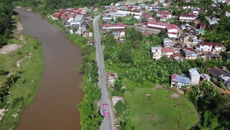 Pink-bus-enters-a-developed-village,-town,-city-beside-a-river-in-the-country-side-of-Madagascar