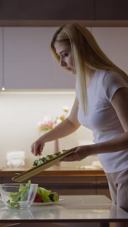 careful blonde woman pours fresh cut cucumbers foe salad into glass bowl from wooden board at table in contemporary kitchen slow motion