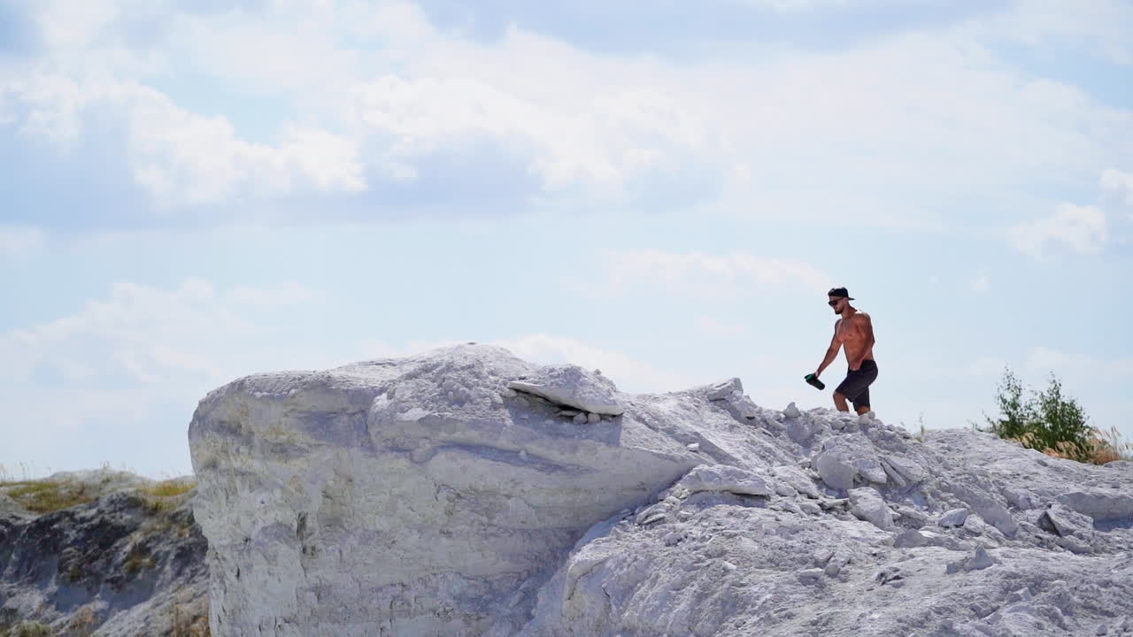 Slow motion of strong athlete in the mountains. Shirtless sportsman walking up the white hill in summer. Muscular man exercises outdoors.