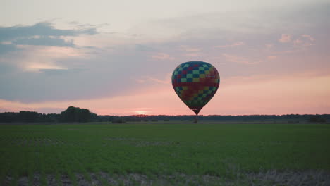 hot air balloon descending towards green field at sunset with checkerboard canopy gliding over vast crop rows under pastel sky horizon creating serene aerial moment filled gentle summer breeze