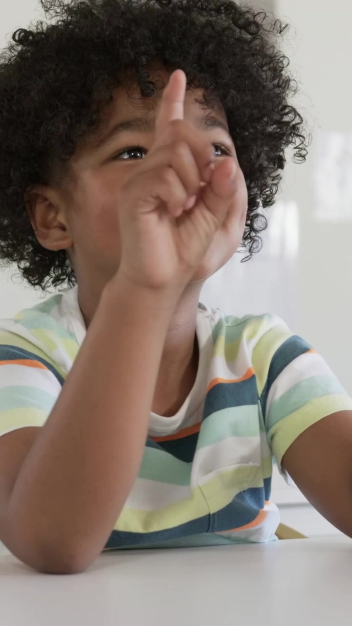 Vertical video: Smiling child sitting at white table wearing striped tee, pointing up from rest