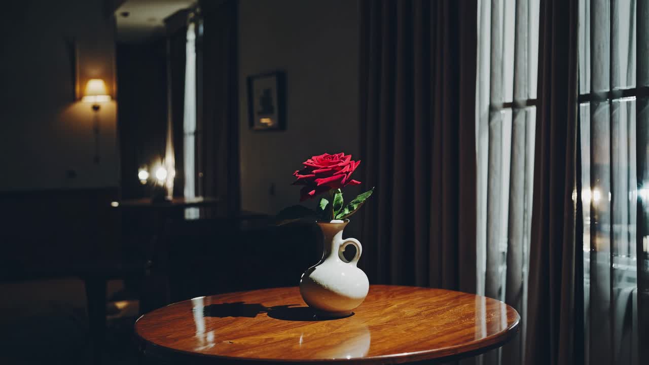 Red rose with green leaves standing in a small white vase placed on a round wooden table in a dark room with a window and curtains slightly open, creating a cozy and intimate atmosphere