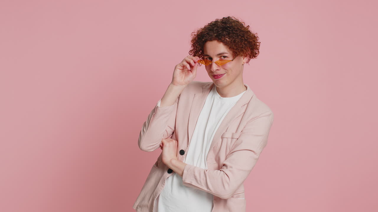 elegante y sonriente joven caucásico con cabello rizado con gafas de sol naranjas expresión alegre