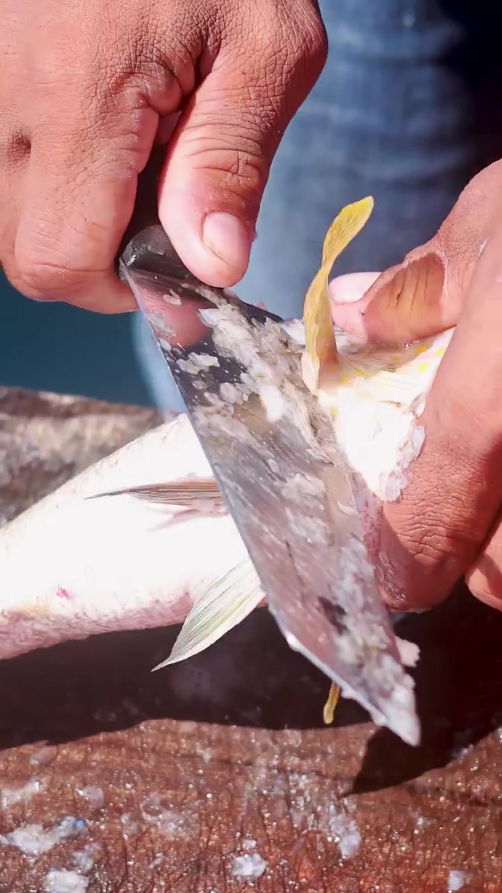 Hands remove scales from a yellowtail fish using a large knife on a wooden surface, under bright natural daylight, with close-up camera angles
