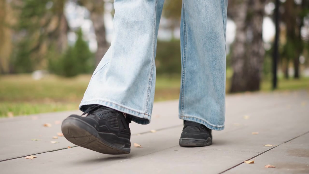 Primo piano delle gambe e dei piedi di una persona che cammina su un marciapiede in un ambiente di parco all'aperto. La persona indossa jeans azzurri a gamba larga e sneakers nere con foglie cadute sparse a terra. Gli alberi sono