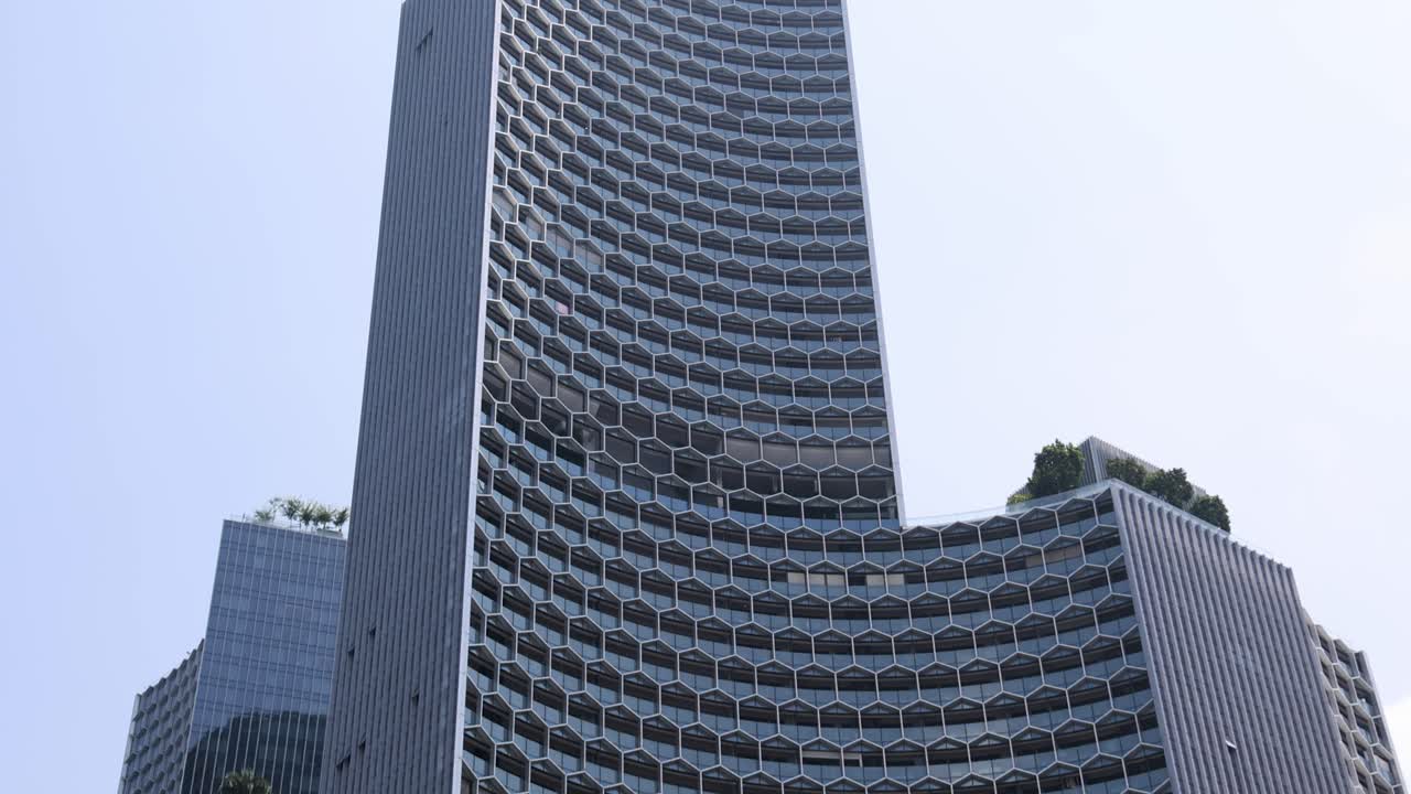 Upward camera tilt reveals modern Singapore high-rise with geometric facade, urban greenery, and bright daylight