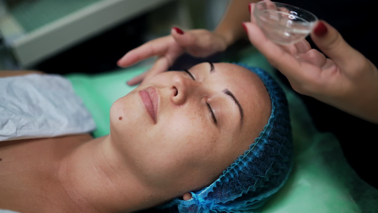 Caucasian woman wearing protective cap lies with eyes closed on the couch at salon. Cosmetologist applying some gel from a little glass cup on client's face. Close up.
