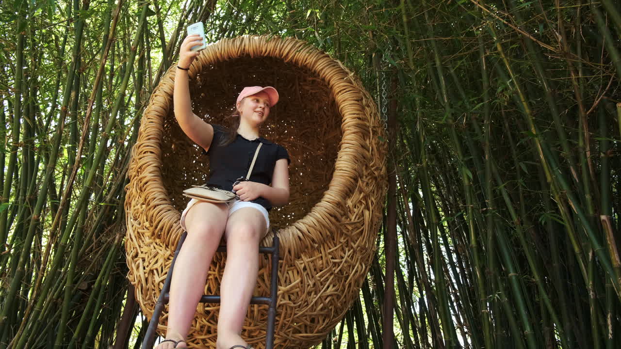 una joven tomando un selfie en un asiento con forma de huevo de mimbre en un bosque de bambú oriental
