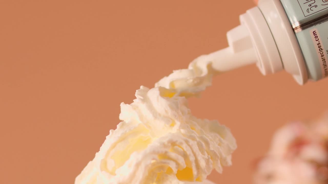 A hand uses an aerosol can to swirl whipped cream onto a vanilla milkshake